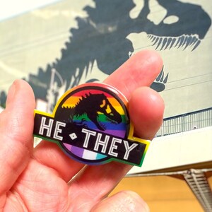 May include: A circular pin featuring a black dinosaur silhouette against a rainbow background. The design includes a black banner at the bottom with the white text HE • THEY. The pin incorporates a pride flag color scheme with stripes of red, orange, yellow, green, blue, and purple, and a smaller section with white, purple, and green stripes.