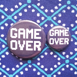 May include: Two round, dark purple buttons with the words "GAME OVER" in white, pixelated font. One button is larger than the other. The buttons are set against a blue fabric background with a white and teal geometric pattern.