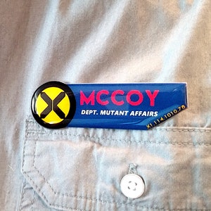 May include: A rectangular blue name badge with a black circular logo featuring a yellow X. The text reads MCCOY and DEPT. MUTANT AFFAIRS in white and red lettering, with the code X1.114.1010.78 printed in the bottom right corner.