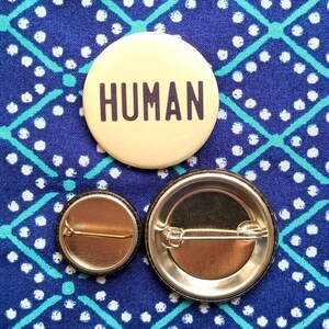May include: A circular pinback badge with a cream-coloured background and the word HUMAN printed in bold black capital letters. Below the badge, two smaller circular pinback badges are shown from the back, revealing metallic pin fasteners.