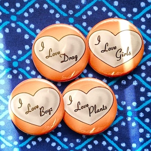 May include: Four orange round buttons with white heart-shaped designs. Each button has the phrase "I Love" followed by "Drag", "Girls", "Boyz", and "Plants" in black script. The buttons are on a blue patterned fabric.