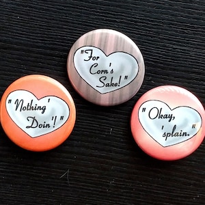 May include: Three heart-shaped button pins with different sayings. The first pin is orange with the text "Nothing' Doin'!". The second pin is brown with the text "For Corn's Sake!". The third pin is pink with the text "Okay, 'splain."