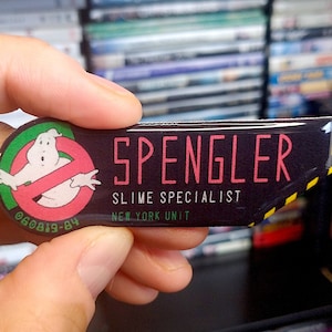 May include: A rectangular pin featuring the Ghostbusters logo and the name "SPENGLER" in pink lettering. The pin also includes the text "SLIME SPECIALIST" and "NEW YORK UNIT" on a black background. The pin has a yellow and black striped border.