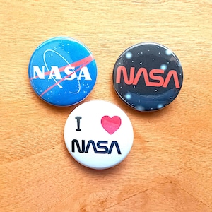 May include: Three round pinback buttons with different NASA designs. The first button is blue with a white NASA logo and a red swoosh. The second button is black with white dots and a red NASA logo. The third button is white with a red heart and the text "I ♥ NASA" in black.