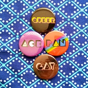 May include: Four circular badges with the words "QUEER", "ACE", "PAN", and "GAY" printed on them. The badges are in various colours, including black, pink, orange, and brown. They are arranged on a blue patterned fabric.