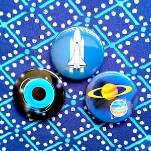 May include: Three circular pinback buttons featuring space-themed designs. One button displays a white space shuttle on a blue background, another shows a black hole with blue rings, and the third depicts a yellow planet with rings and a smaller blue planet.