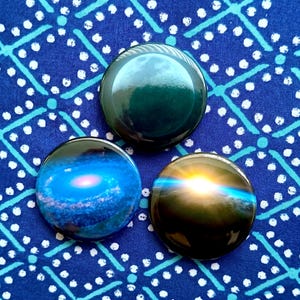 May include: Three round glass magnets with celestial designs. One is a deep green, the second depicts a blue galaxy with a pink center, and the third shows a sunset with a blue streak. They rest on a blue patterned fabric.