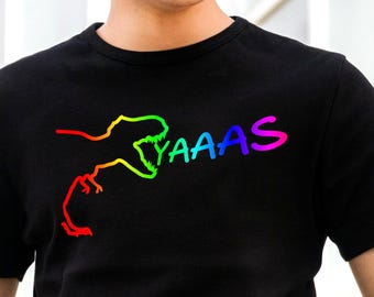 May include: Black t-shirt featuring a rainbow-colored dinosaur graphic with the word "YAAAS" in a matching rainbow font. The design is a playful and colorful statement piece.
