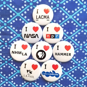 Peut inclure: Huit badges ronds blancs disposés en forme de pyramide. Chaque badge présente le texte I, un symbole de cœur rouge et le nom ou logo d'une organisation spécifique, notamment LACMA, NASA, NPR, NHMLA, PBS, HAMMER, LA PHIL et California Science Center.