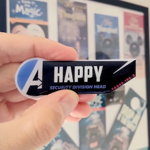 May include: A rectangular black and blue name badge featuring a white letter A logo on the left side. The text HAPPY is printed in large white capital letters, with SECURITY DIVISION HEAD below it and the red code T0S40-196-3 on the right edge.