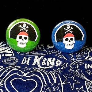 May include: Two round pirate-themed buttons. One is green, the other blue. Each features a white skull wearing a red bandana, with crossed bones on a black background. The buttons rest on a blue fabric with the words "BE KIND".