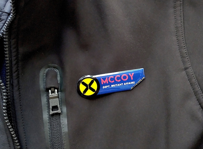 May include: A rectangular name badge with a rounded left side attached to a black jacket. The badge features a yellow circle with a black X, the name MCCOY in pink, and the text DEPT. MUTANT AFFAIRS in white on a blue background.