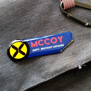 May include: A rectangular blue enamel pin with a rounded left side featuring a yellow circle and black X symbol. The text MCCOY is printed in red, with DEPT. MUTANT AFFAIRS and the code X1.114.1010.78 in white lettering on the right side.