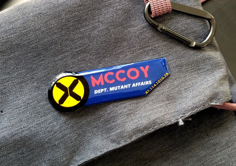 May include: A rectangular blue enamel pin with a rounded left side featuring a yellow circle and black X symbol. The text MCCOY is printed in red, with DEPT. MUTANT AFFAIRS and the code X1.114.1010.78 in white lettering on the right side.