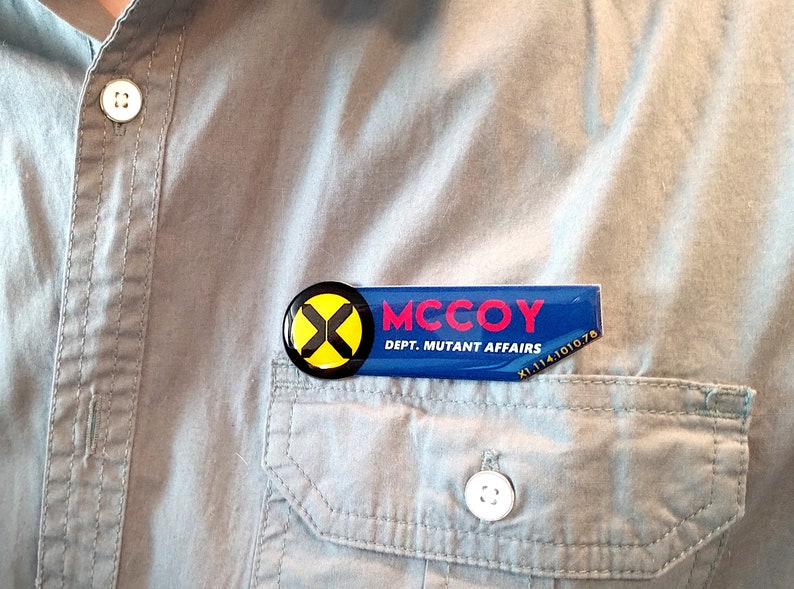 May include: A rectangular blue name badge with a black circular logo featuring a yellow X. The text reads MCCOY and DEPT. MUTANT AFFAIRS in white and red lettering, with the code X1.114.1010.78 printed in the bottom right corner.