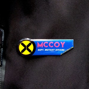 May include: A rectangular blue name tag pin featuring a yellow circle with a black X logo on the left side. The text MCCOY is printed in red, with DEPT. MUTANT AFFAIRS and X1.114.1010.78 printed in white on a black fabric background.