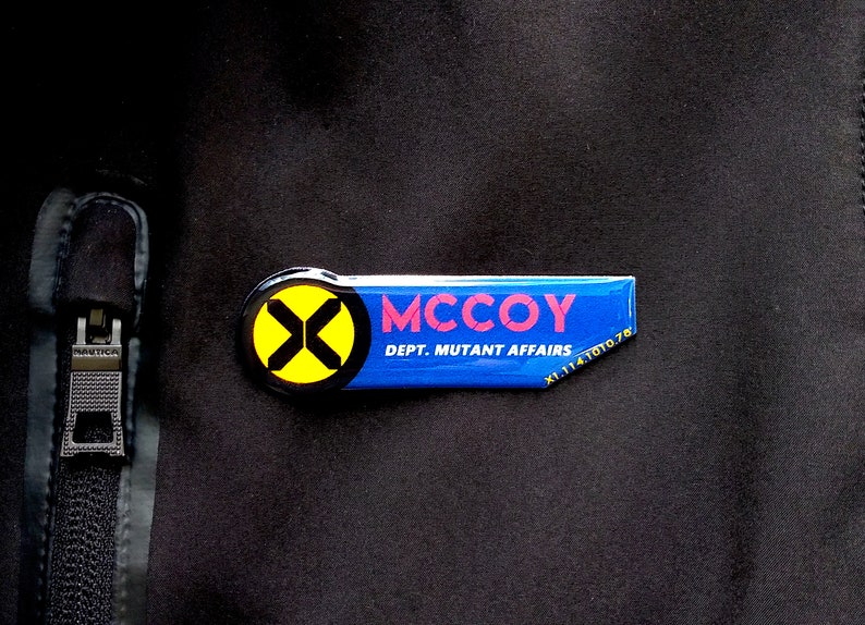 May include: A rectangular blue name tag pin featuring a yellow circle with a black X logo on the left side. The text MCCOY is printed in red, with DEPT. MUTANT AFFAIRS and X1.114.1010.78 printed in white on a black fabric background.