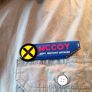 May include: A rectangular name badge with a circular yellow and black X logo on the left. The blue badge features the red text MCCOY and the white text DEPT. MUTANT AFFAIRS, with the small white alphanumeric code X1.114.1010.78 in the corner.