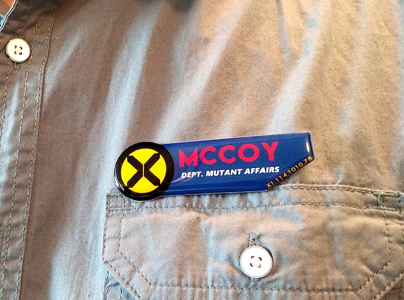 May include: A rectangular name badge with a circular yellow and black X logo on the left. The blue badge features the red text MCCOY and the white text DEPT. MUTANT AFFAIRS, with the small white alphanumeric code X1.114.1010.78 in the corner.