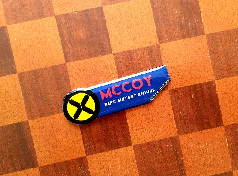 May include: A blue and yellow badge with the text 'MCCOY DEPT. MUTANT AFFAIRS' and a yellow X with black lines on a blue background.