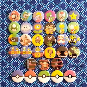 May include: A collection of colorful button pins featuring various characters and elements from the Super Mario and Pokemon franchises. The pins include images of Mario, Luigi, Princess Peach, Yoshi, and Poke Balls.