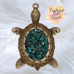 Op de afbeelding: Een gouden schildpadbedel met een groene glitter epoxy vulling. De schildpad heeft een gedetailleerde schild en poten. De bedel is gelabeld "Cute Nyss by Enayah".