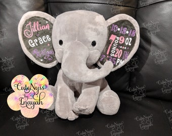 Op de afbeelding: Een grijze pluche olifant met roze en paarse geborduurde oren. Het ene oor zegt "Jillian Grace" en het andere oor zegt "10.18.18 7 pond 9 oz 20 inch 7:35 PM".