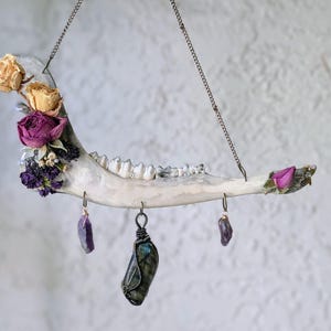 May include: A hanging animal jawbone decorated with dried yellow and purple roses, small white and purple flowers, and three dangling crystals. A large wire-wrapped labradorite stone hangs in the center, flanked by two smaller purple amethyst-like crystals, all suspended by a thin metal chain.