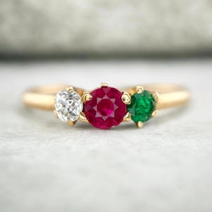 May include: A gold ring featuring three round-cut gemstones set in a row. The center stone is a vibrant red ruby, flanked by a clear white diamond on the left and a deep green emerald on the right, all secured by small gold prongs on a smooth band.