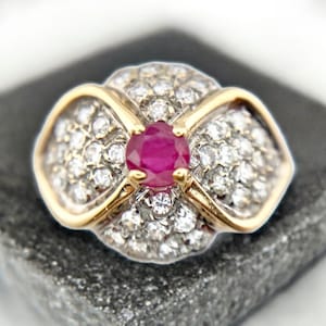 May include: A gold-toned ring with a floral design. The center features a round, vibrant red gemstone, surrounded by a cluster of small, clear gemstones. The ring is set on a dark gray surface.
