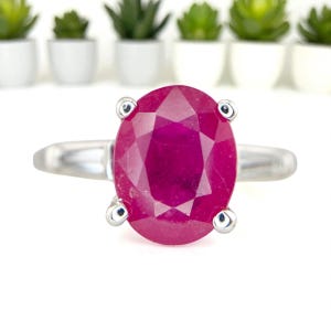 May include: A silver ring featuring a large, oval-cut ruby gemstone. The ruby is a vibrant shade of red and is held in place by four silver prongs. The ring's band is a simple, polished silver.