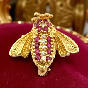 Puede incluir: Broche dorada con forma de abeja con detalles de rubíes y diamantes. El cuerpo de la abeja está adornado con pequeños rubíes redondos y una fila central de diamantes. Las alas están detalladas con líneas grabadas.