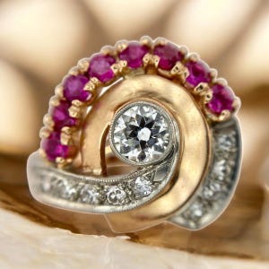 Peut inclure: Une bague vintage avec un diamant central étincelant, entouré d'un tourbillon d'or et d'argent. Une rangée de rubis rouges vifs forme une arche au-dessus, avec de petits diamants sur la bande. La bague est posée sur une surface neutre.