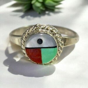 May include: A silver ring with a round, inlaid stone. The stone is divided into four sections: white, black, red, and green. The ring has a braided border around the stone.
