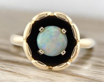 May include: A gold ring featuring a round, iridescent opal gemstone set in a black, flower-shaped setting. The ring has a delicate, floral design around the opal, and a simple gold band. The opal displays a range of colors, including blue, green, and white.