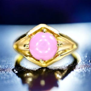 May include: A gold ring with a large, round, pink gemstone set in the center. The ring has a simple, elegant design.