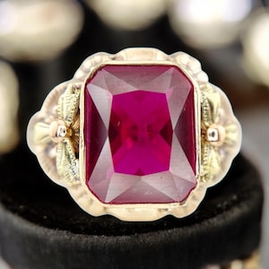 Puede incluir: Un anillo de oro con una gran gema rectangular de color rojo intenso. El anillo tiene un engaste ornamentado con detalles florales y de hojas. La gema es el punto focal, mostrando su rico color. Una pieza de joyería llamativa.