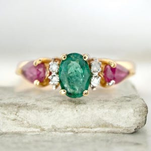 Puede incluir: Un anillo de oro con una esmeralda verde central de corte ovalado, flanqueada por dos rubíes rosas de corte en forma de pera. La esmeralda está acentuada por dos pequeños diamantes blancos de corte redondo a cada lado, en una banda de oro pulido.