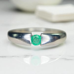 Puede incluir: Un anillo de plata con una esmeralda verde brillante en el centro. El anillo tiene un diseño moderno y minimalista con un acabado pulido. La esmeralda es redonda y vibrante, engastada entre dos secciones elevadas de la banda.