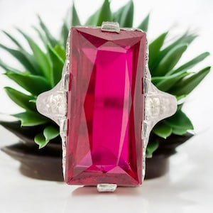 May include: A silver-toned ring featuring a large, rectangular, faceted, magenta-colored gemstone. The ring has ornate detailing on the band and around the gemstone setting. The ring is displayed against a backdrop of green foliage.