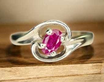 May include: A silver ring featuring a vibrant oval-cut ruby gemstone. The ruby is set in a swirling, openwork design. The ring's band has a smooth, polished finish, creating a classic and elegant look. The ring is displayed on a wooden surface.