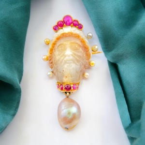 Puede incluir: Broche antiguo detallado con un rostro tallado, adornado con piedras preciosas rosas y perlas. El broche tiene un engaste de oro y un gran colgante de perla en forma de pera. El fondo incluye una tela verde azulado.