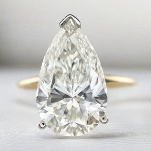 May include: A pear-cut diamond ring featuring a large, faceted, colourless stone set in a three-prong silver-toned metal setting. The ring band is crafted from a smooth, polished yellow gold metal, creating a contrast with the silver prongs.