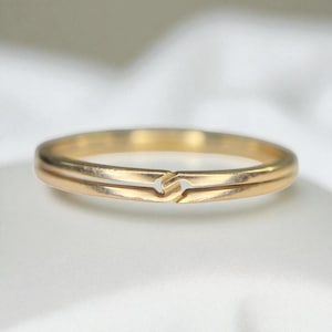 May include: A gold-colored ring with a unique design. The ring features a split band that connects at the front, creating an interesting visual detail. The ring is set against a soft, white background.