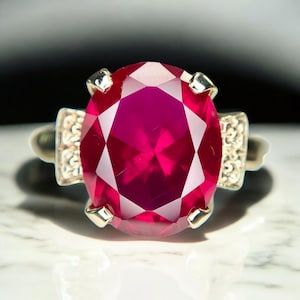 May include: A close-up of a gold ring featuring a large, oval-cut ruby gemstone. The ruby is a vibrant shade of red with multiple facets. The ring has decorative silver accents on either side of the ruby.