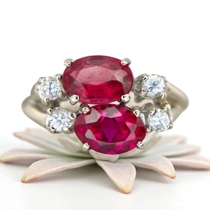 May include: A silver ring featuring two large oval-cut, deep red rubies and four smaller, round, clear gemstones. The ring is set on a light pink and white flower.