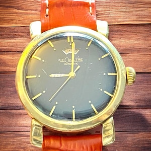 May include: A gold-tone wristwatch with a black face and a brown leather band. The watch face has gold-tone hands and hour markers, and the text "LE COULTRE AUTOMATIC" is printed on the face.