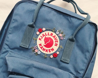 May include: A blue Fjällräven Kånken backpack with a white embroidered patch featuring a fox and the words "Fjällräven Kånken". The patch is surrounded by a wreath of embroidered flowers in pink, red, and white.