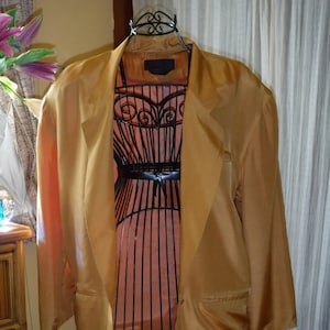 May include: A gold satin blazer with a long, flowing silhouette. The blazer has a single button closure and two pockets. It is displayed on a black wire mannequin.