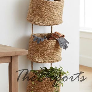 Puede incluir: Tres cestas redondas de yute tejido en un tono marrón natural, colgadas de una pared. La cesta superior contiene papeles y llaves, la del medio tiene guantes y la inferior una planta verde. Zapatillas blancas en la parte inferior.